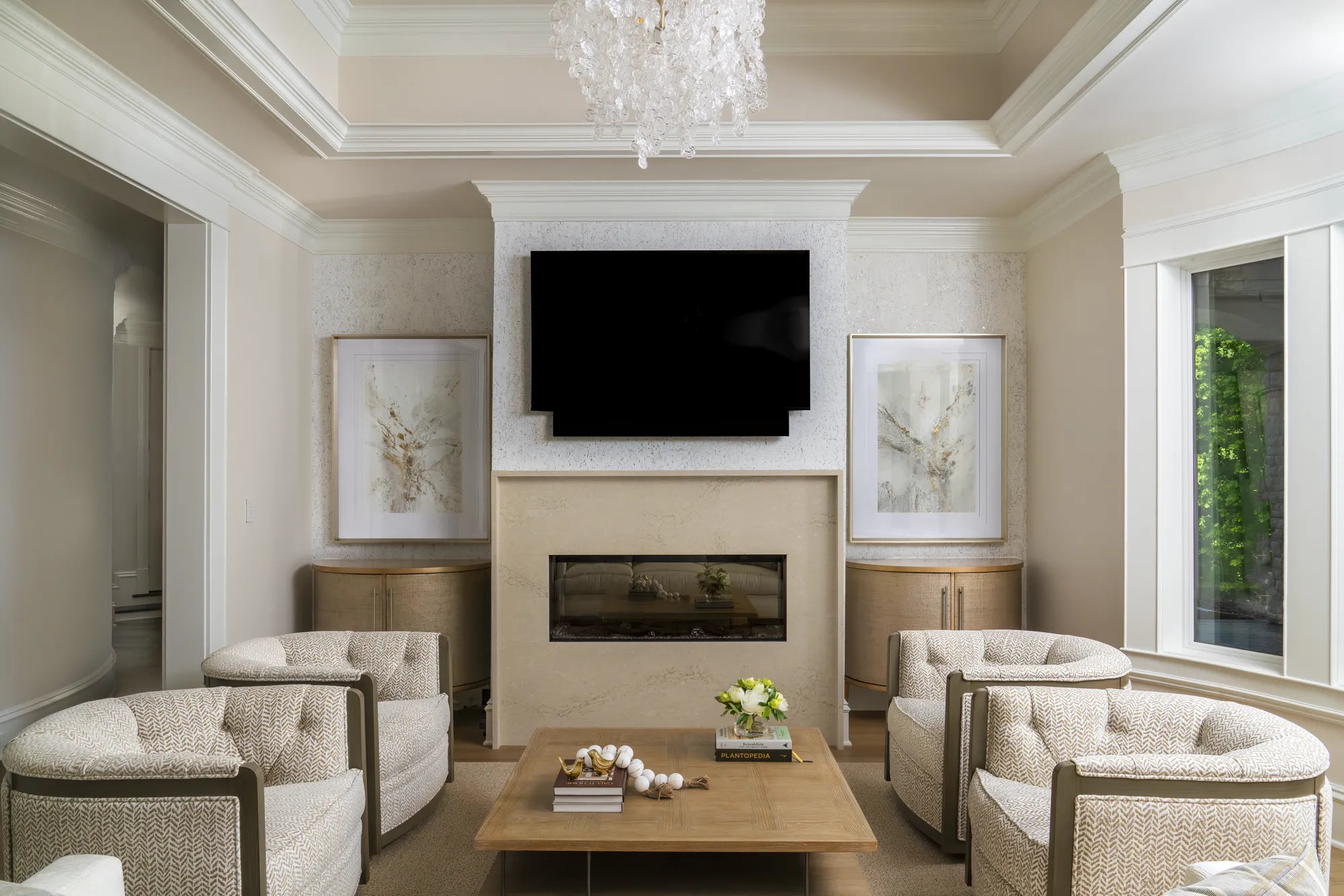 A sophisticated living space with cream-colored seating arranged around a wood coffee table. The neutral room features crown molding, a modern chandelier, and a mounted TV above a light stone fireplace.