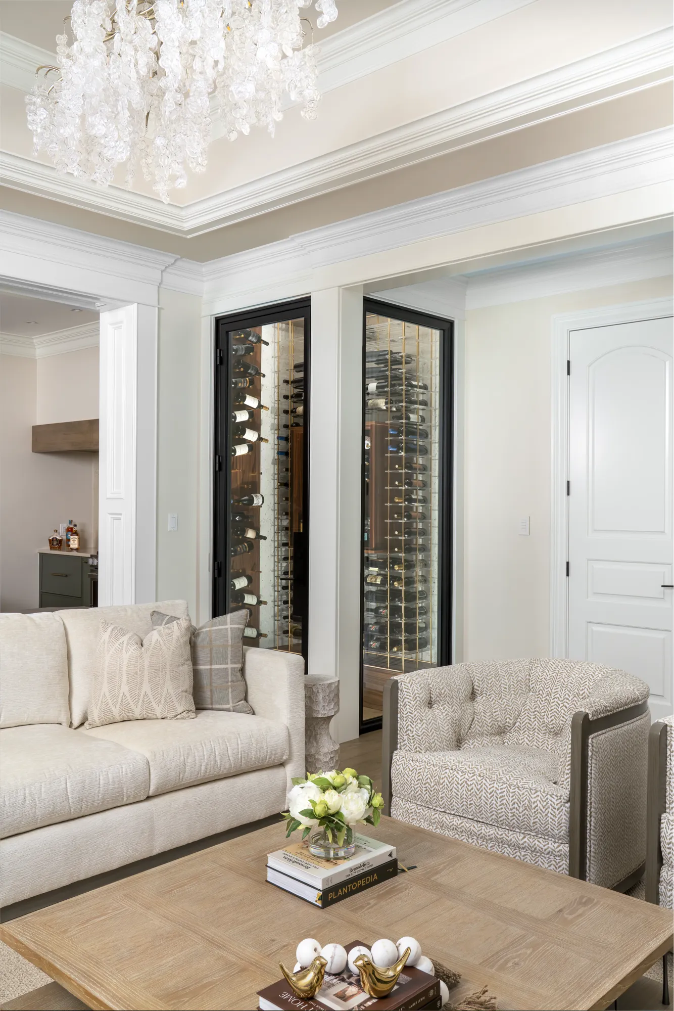 A luxurious living space featuring a tall, glass-fronted wine display built into the wall with brass racking holding many bottles. Neutral sofa and armchair sit opposite a wooden coffee table.