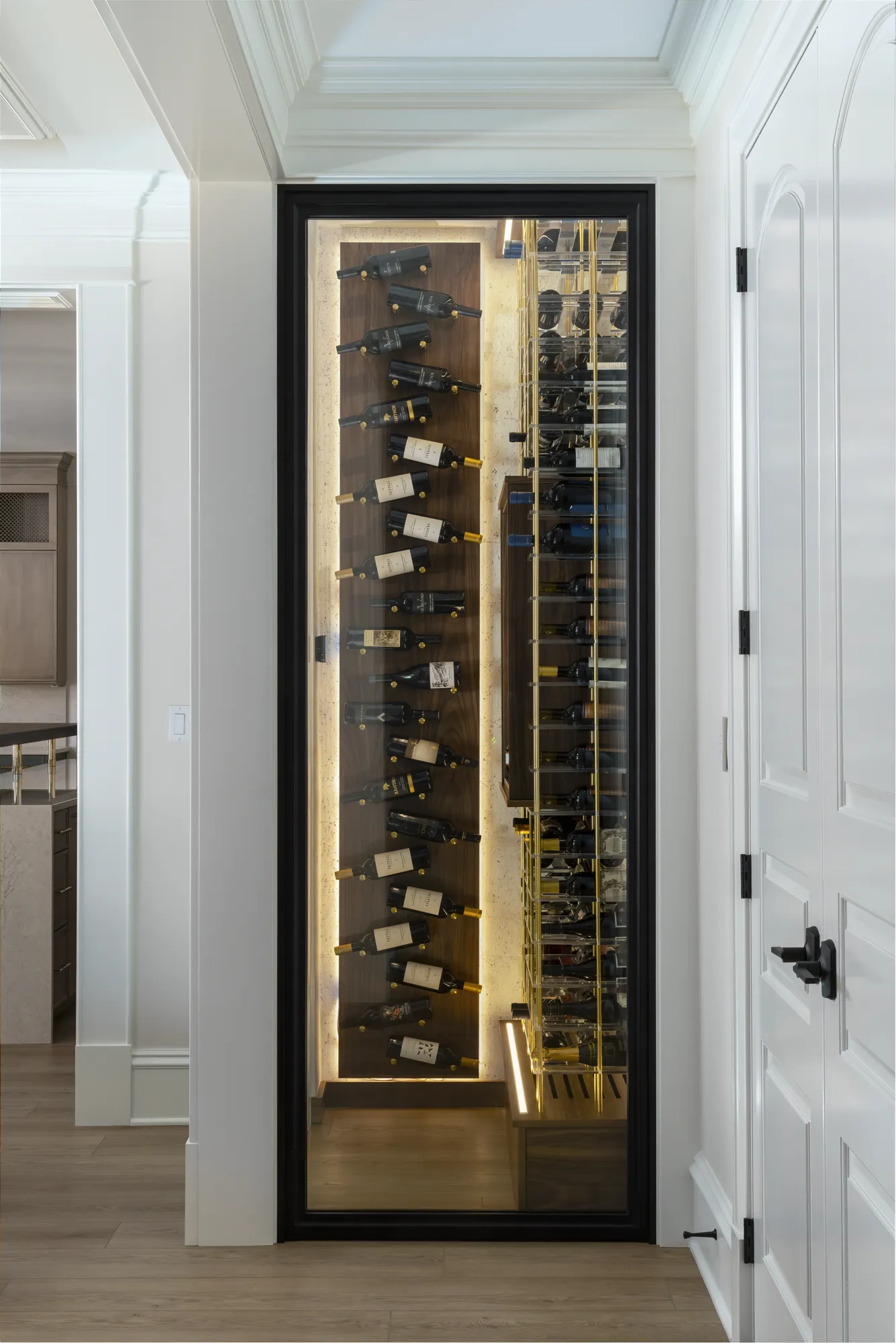A narrow, modern wine display built into a hallway wall between a white paneled door and another room. The lighted glass case features floating bottles on a wood panel and gold grid racks.