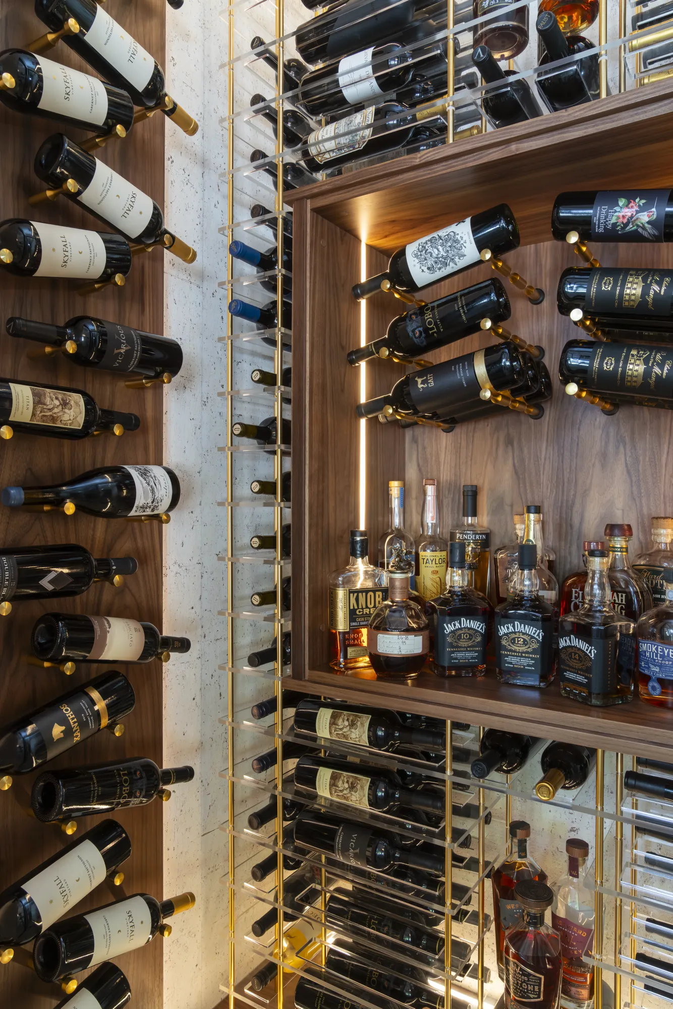 An illuminated, custom liquor and wine storage system features dark wood, clear acrylic shelving, and gold-accented bottle racks.